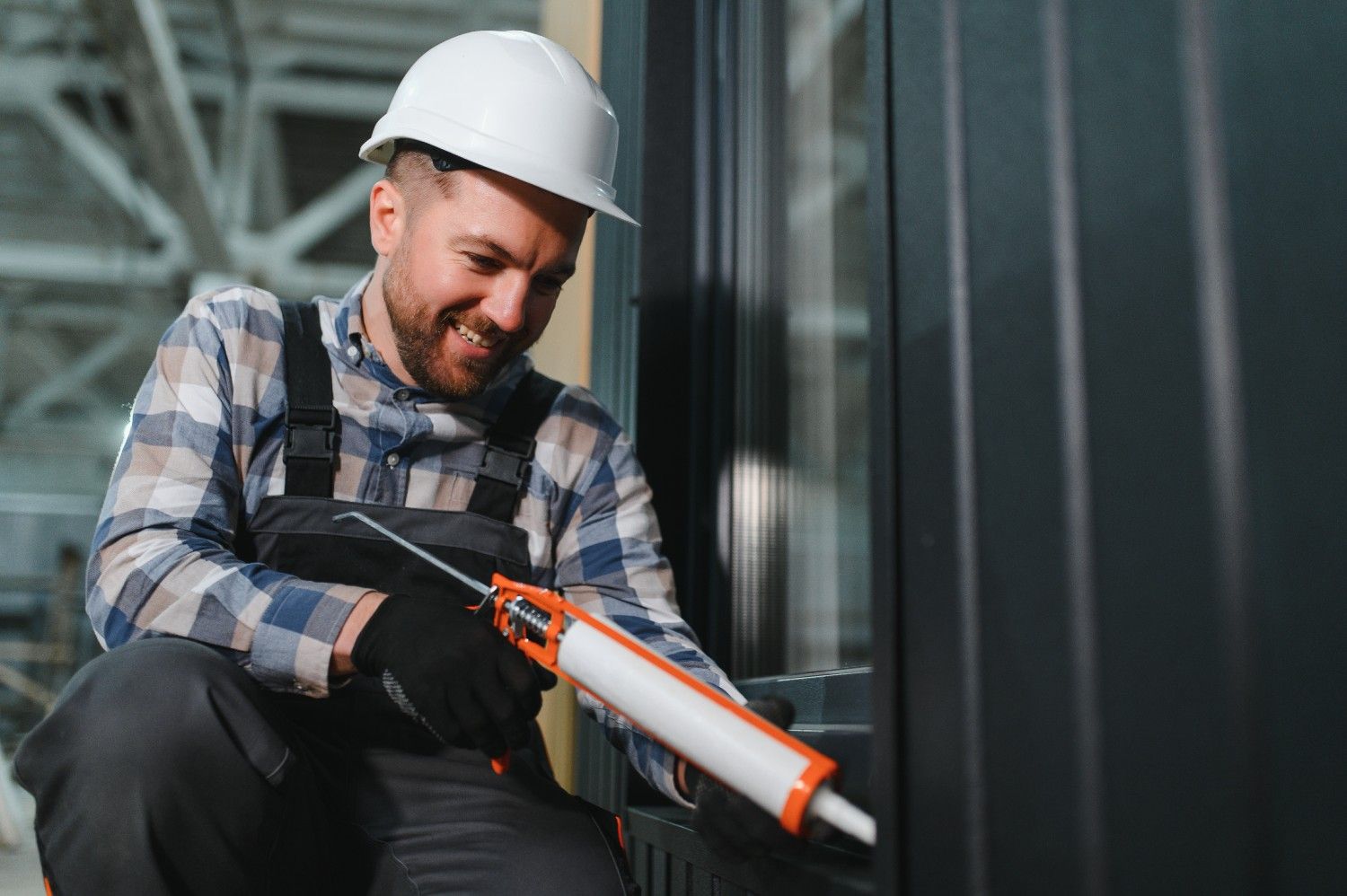 Smiling worker in a hard hat applying sealant with a caulking gun to a dark window frame